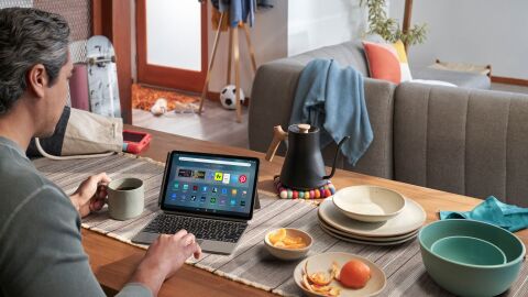 looking over a man's shoulder as he sits at a dining table covered in plates, a kettle, and table runner. he is browsing his fire max 11 tablet with a keyboard accessory