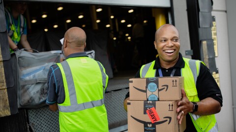 A smiling man holds boxes while people in the background load a truck. 
