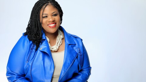 An image of a woman sitting on a chair, smiling for a photo against a white background while wearing a bright blue blazer.