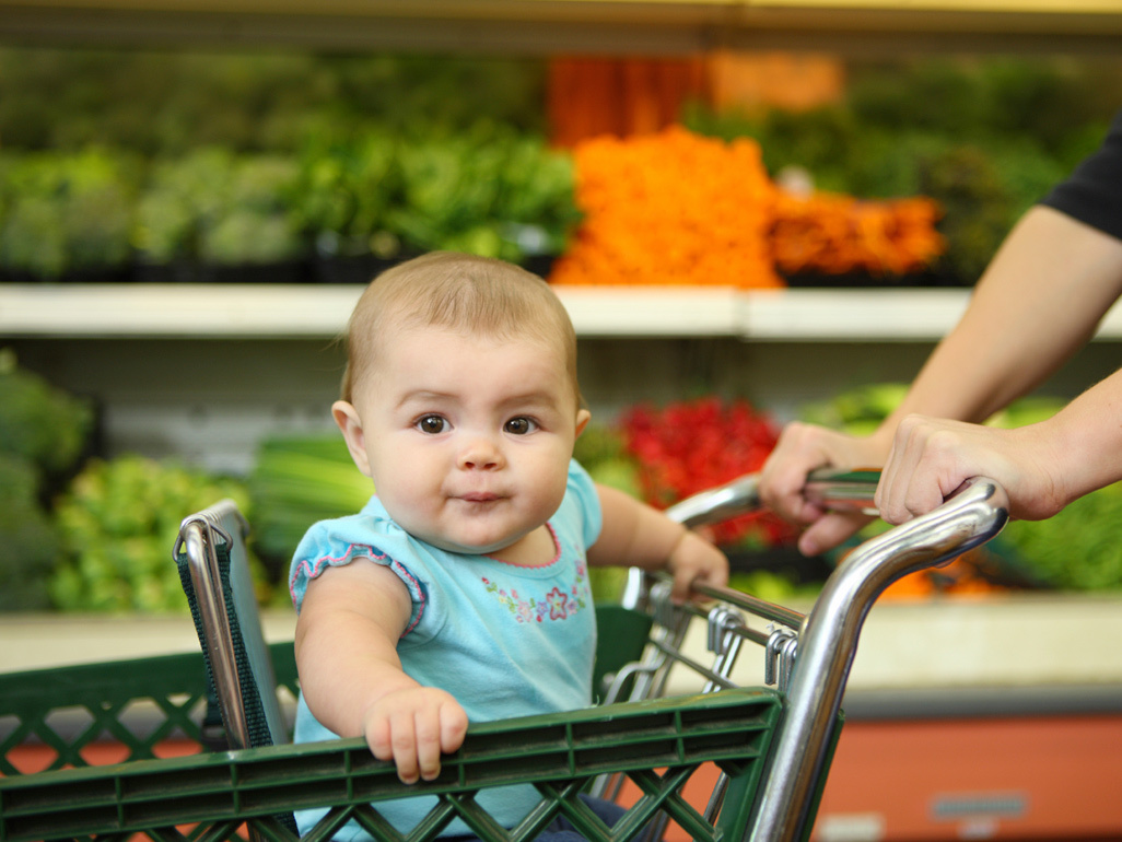 A baby sitting in a shopping cart seat in front of a produce aisle