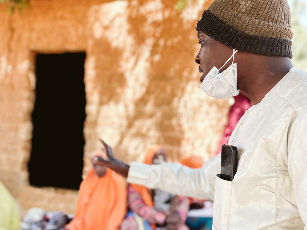 A man stands and speaks to a group of people seated on the ground near a building in Kano State, Nigeria.