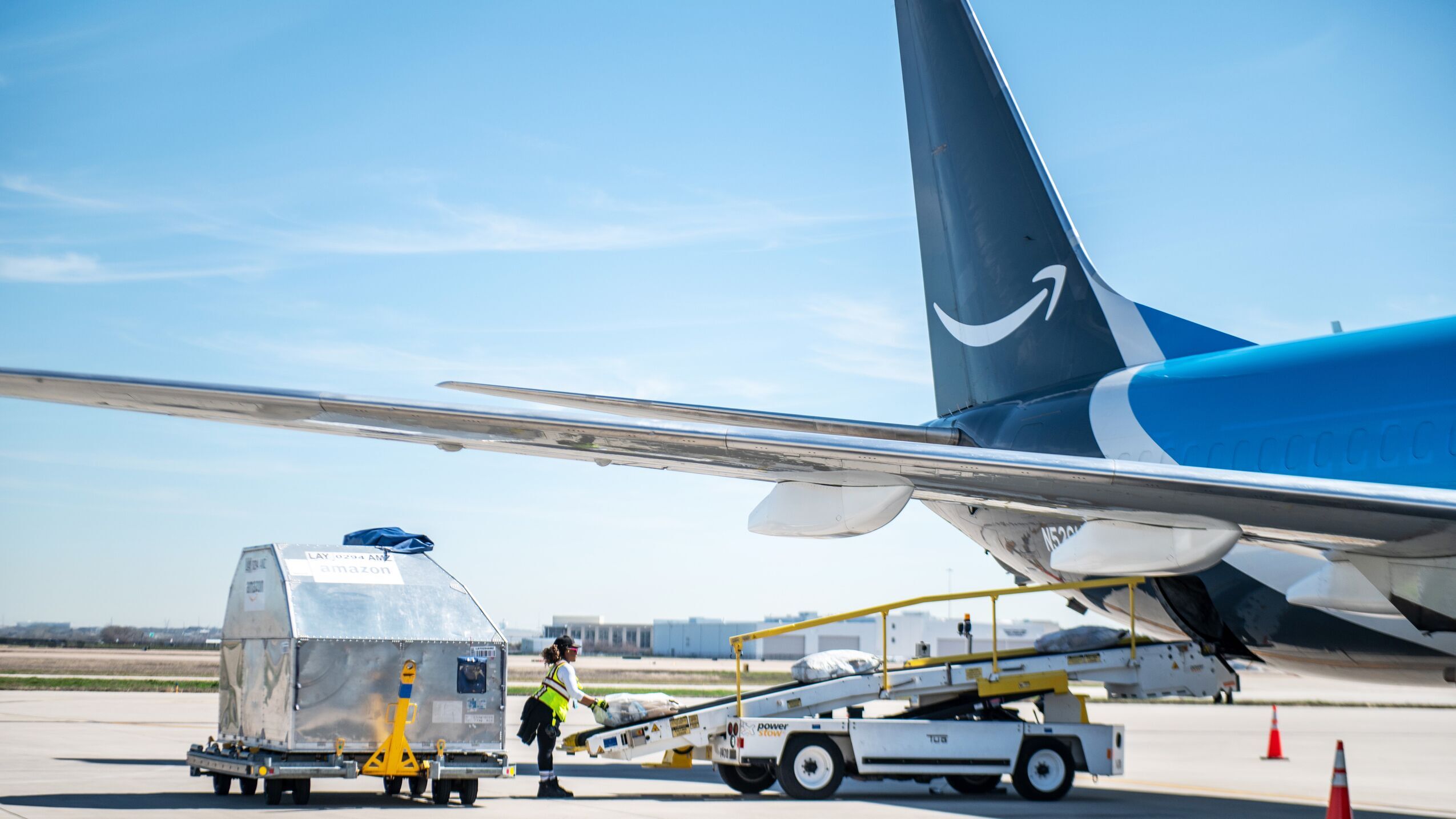 An image of an Amazon Air plane parked outside of the Air Hub. The back of the plane is featured in this image, showing a car pulled up to the plane and using a mobile conveyer belt to load large bags into the belly of the plane.