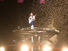 Angus, aged 55, stand atop a large, high pedestal. He is bare-chested and without his cap, he wears shorts while playing his guitar.