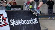 A skateboarder balances at the top of a ramp, which has the Skateboard GB logo on it.