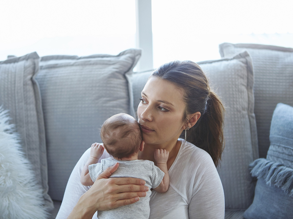 Mum on blue sofa holding new baby and looking distractedly into distance