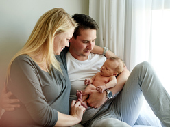 new parents sitting on a bed next to the window with their newborn in father’s arms