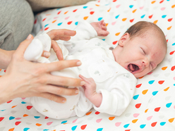 Crying baby lying on a printed sheet while her parent holds her legs to soothe her