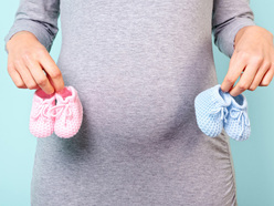 Mulher branca grávida, de vestido cinza, segura um sapato azul e um sapato rosa em frente à barriga 