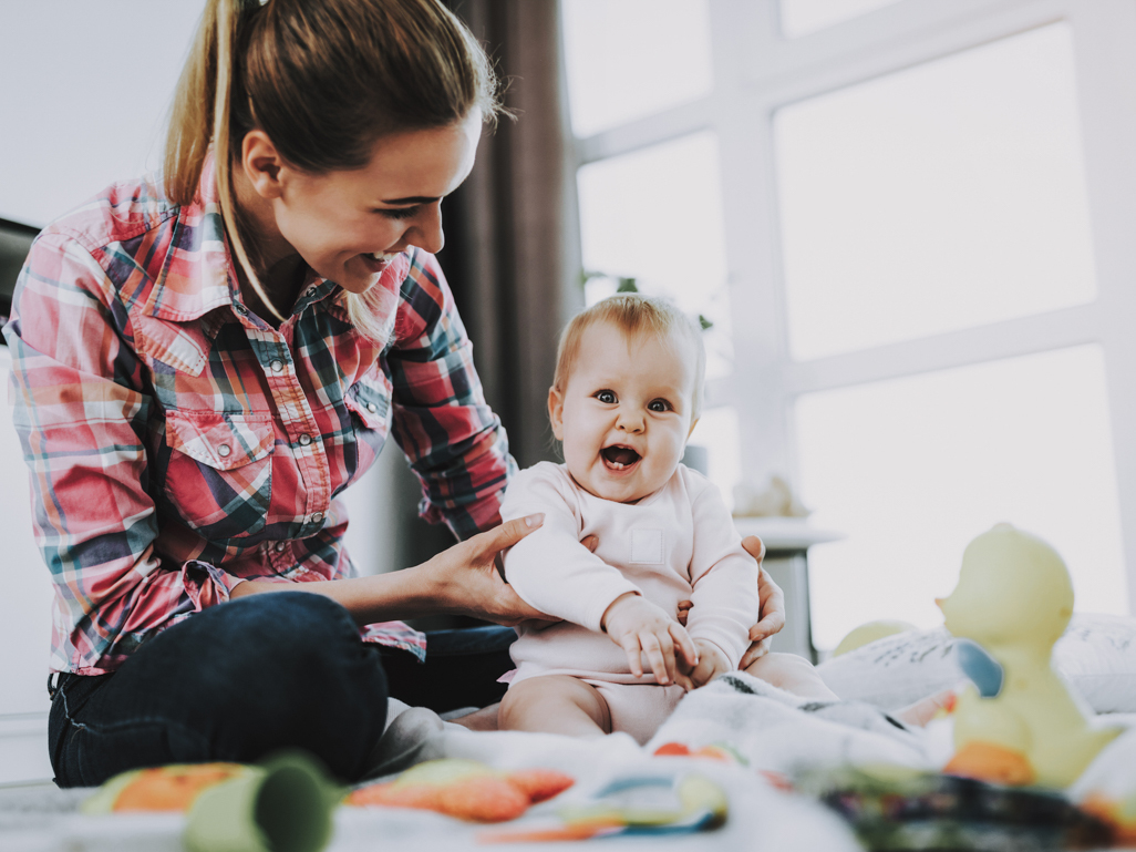 A mom sitting on the floor with her infant, who's smiling at the camera.