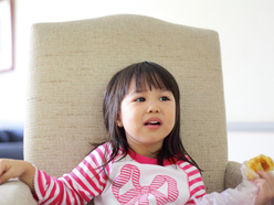 asian girl sitting in a big chair and holding some solid food in her left hand while trying to tell something