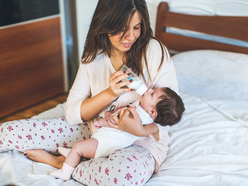 A mom in her pajamas sitting on a bed, bottle-feeding her newborn baby.