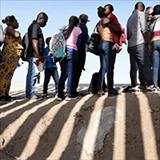 A diverse group of men, women and children stand in line at the border