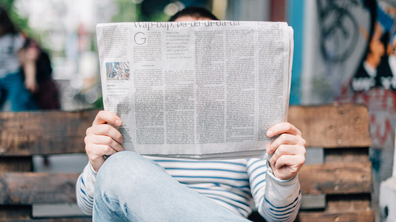 Man sitting on a bench holding a newspaper that is covering his face 