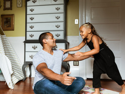 Dad sitting on sitting room floor with his arms outstretched towards young girl