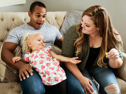 Young girl leaning back on her dad's knee on the sofa and holding her hand out to her mom